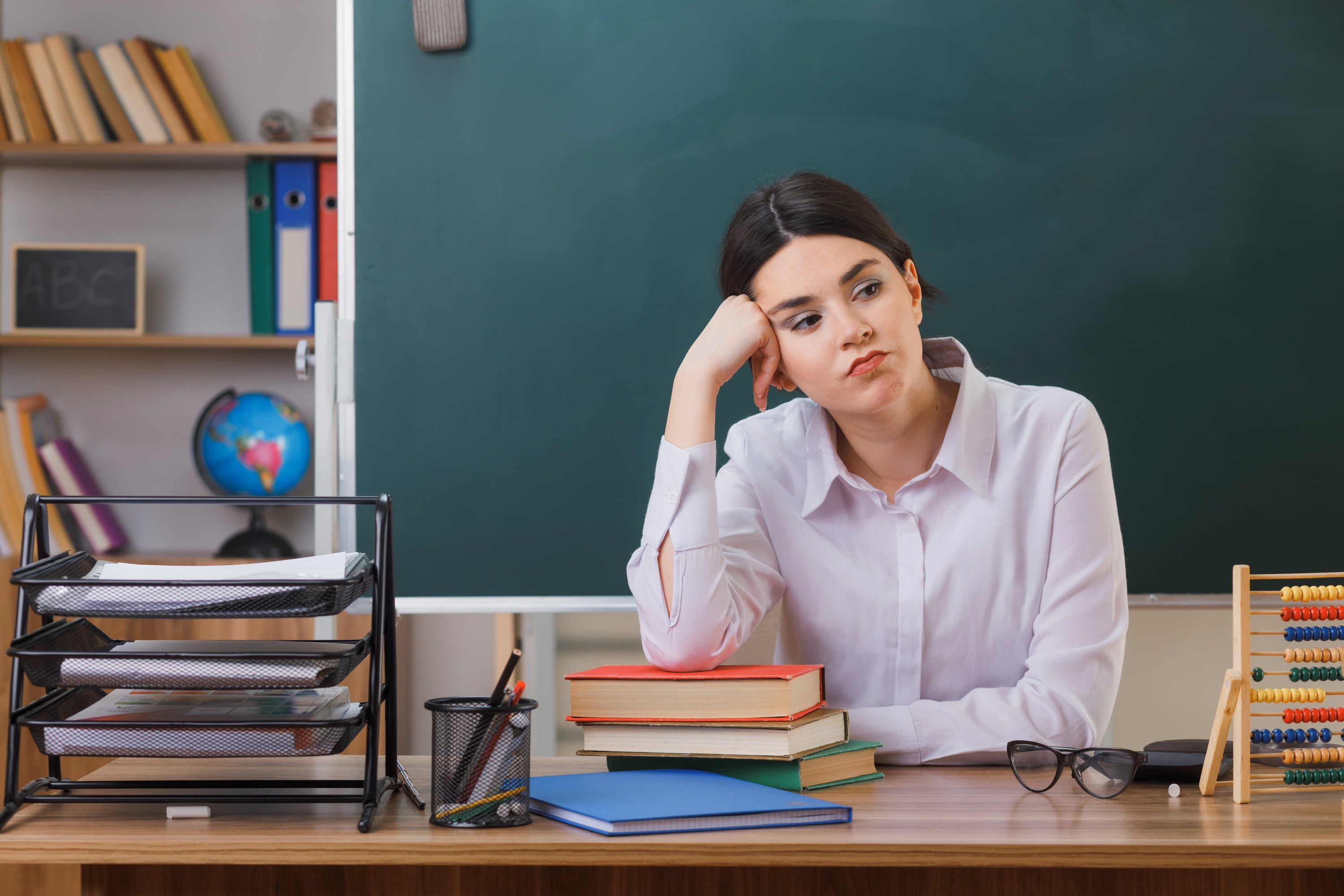 tired putting hand on cheek young female teacher sitting at desk with school tools in classroom
